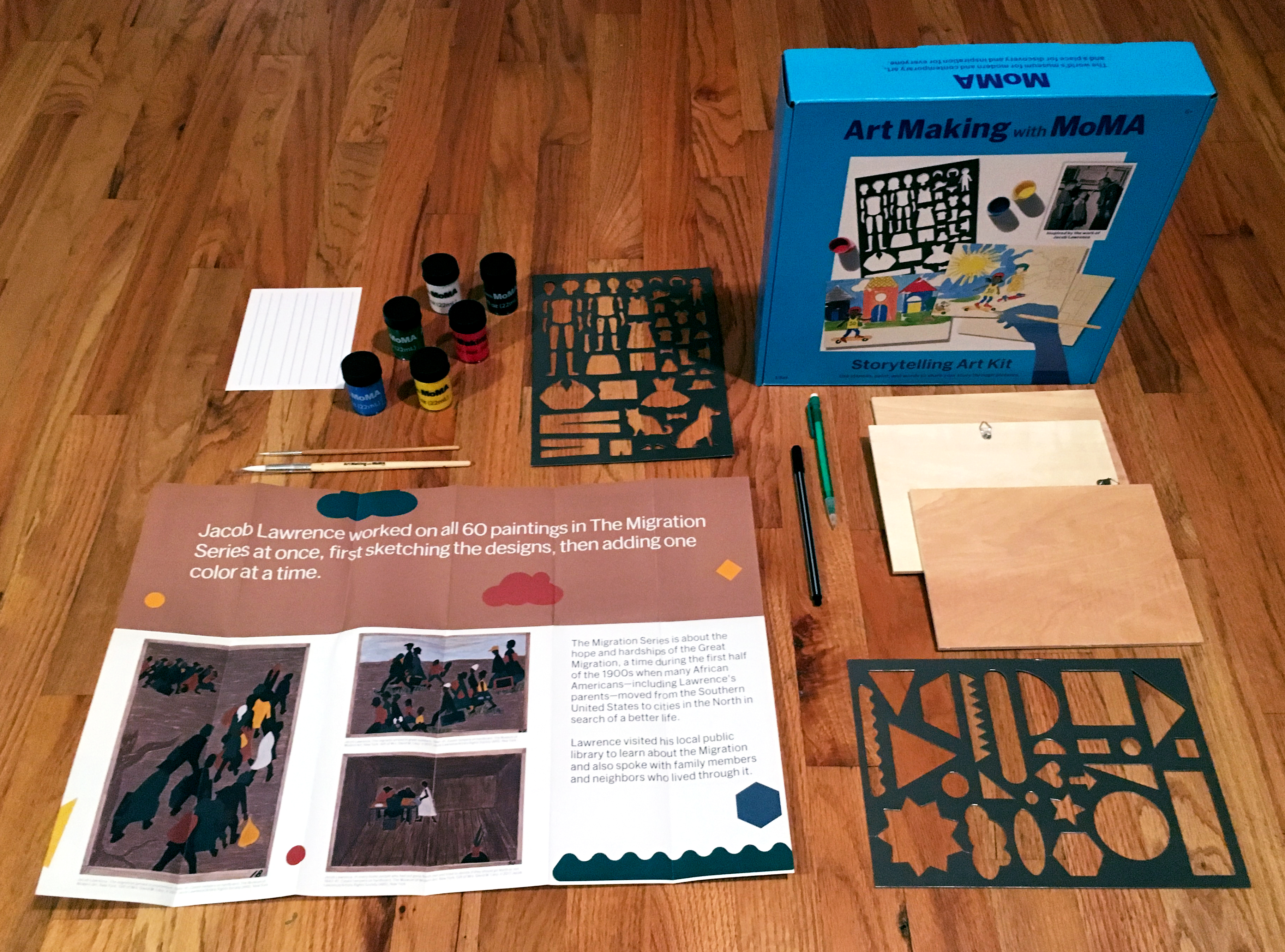 The contents of the Jacob Lawrence storytelling kit neatly laid out on the floor includes a pamphlet, paints, stencils, paintbrushes, and three wooden boards.